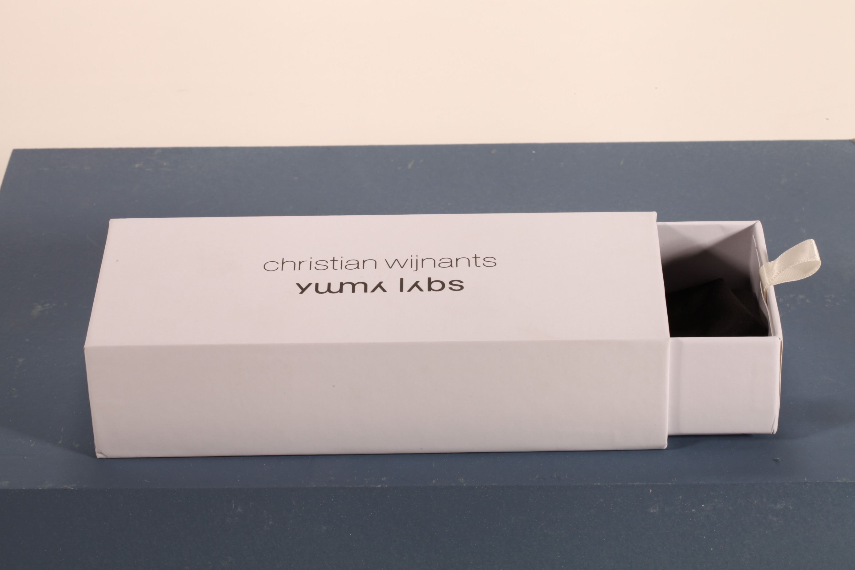 Here is a 50-word English description for the eyewear set shown in the image: This eyewear set features a white paper box and matching soft pouch, measuring 17×4×7 cm. Branded with Christian Wijnants