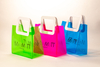 Three translucent bags in green, pink, and blue feature white square handles. The front displays the black text "MARMOTT EYEWEAR".