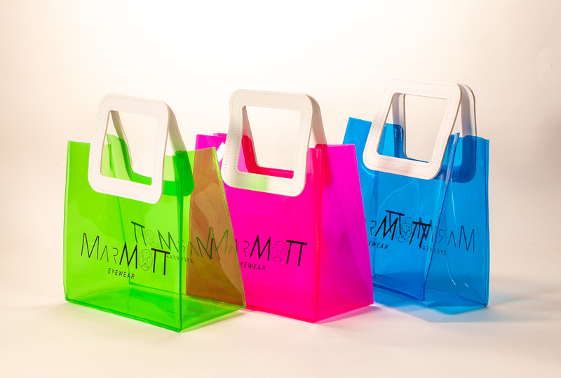 Three translucent bags in green, pink, and blue feature white square handles. The front displays the black text "MARMOTT EYEWEAR".