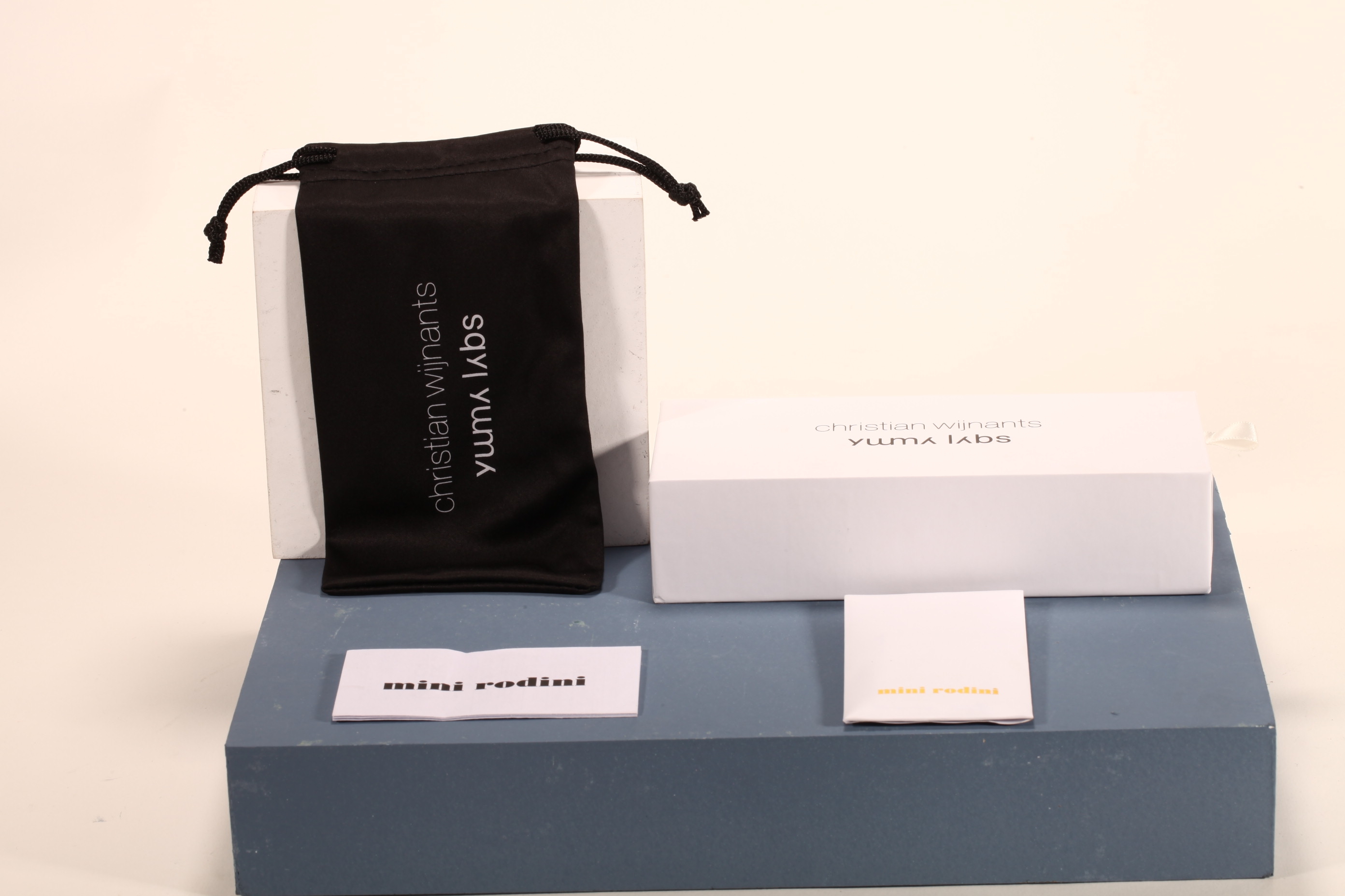 Here is a 50-word English description for the eyewear set shown in the image: This eyewear set features a white paper box and matching soft pouch, measuring 17×4×7 cm. Branded with Christian Wijnants