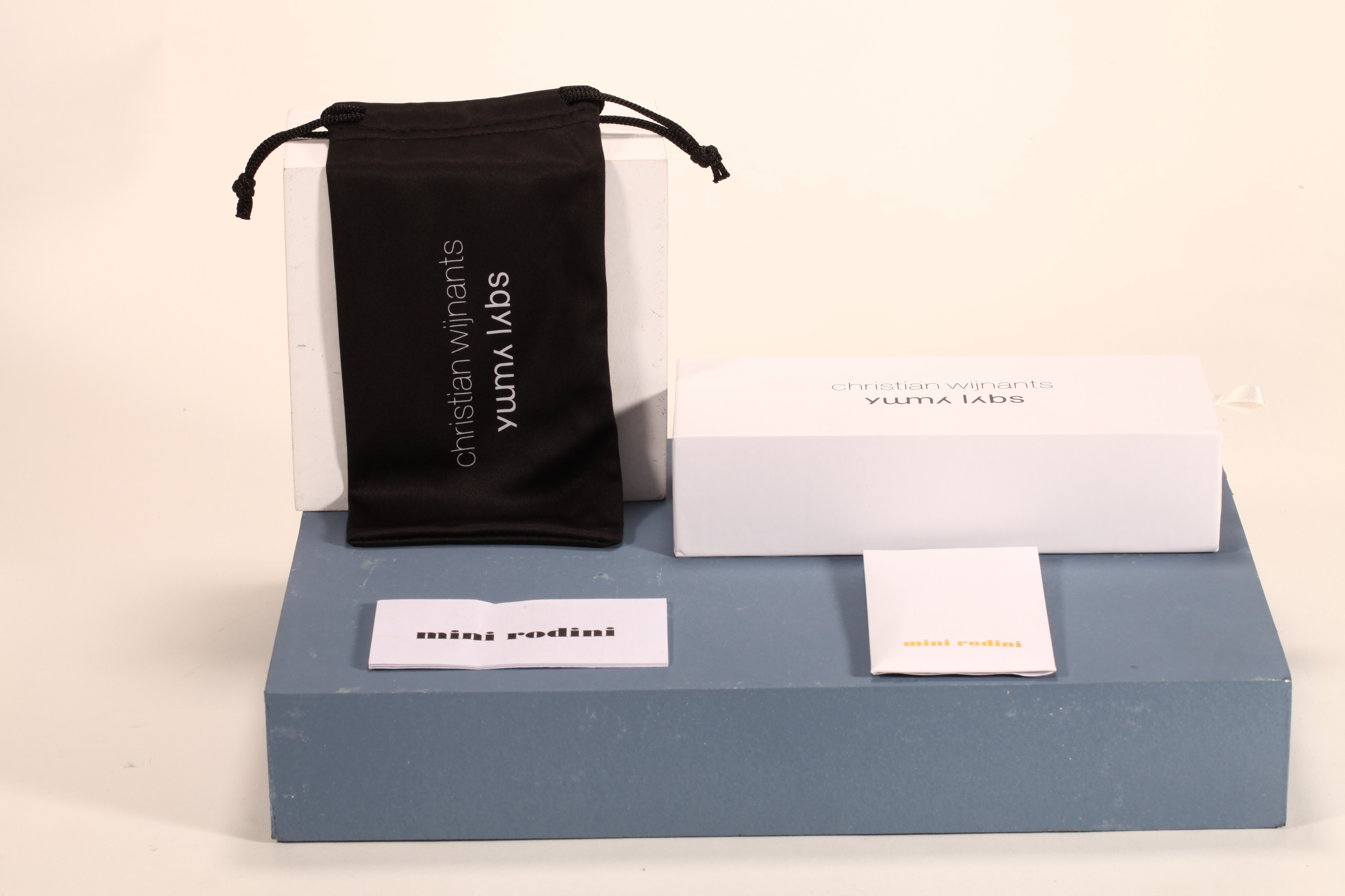 Here is a 50-word English description for the eyewear set shown in the image: This eyewear set features a white paper box and matching soft pouch, measuring 17×4×7 cm. Branded with Christian Wijnants
