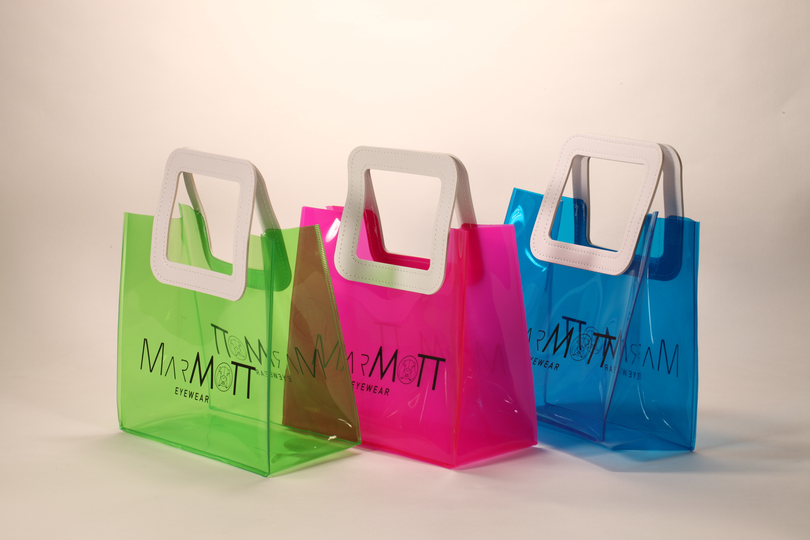 Three translucent bags in green, pink, and blue feature white square handles. The front displays the black text "MARMOTT EYEWEAR".